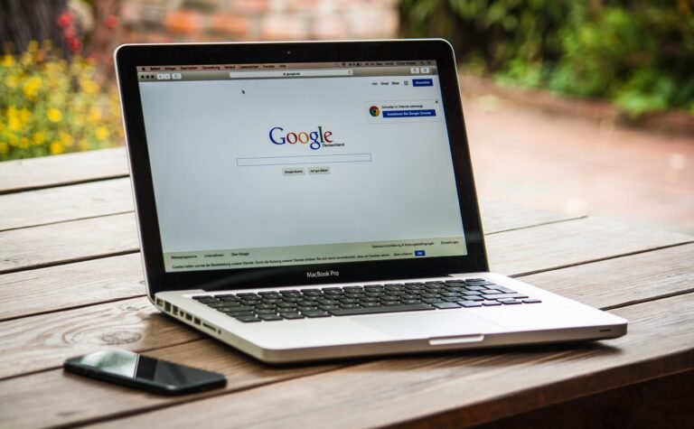 Panduan SEO untuk Pemilik Bisnis: Cara Mendapatkan Pelanggan dari Google A MacBook Pro displaying Google Search on a wooden table outdoors, next to a smartphone.